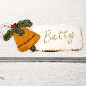 May include: Handmade Christmas ornament featuring a felt bell and a tag embroidered with the name "Betty". The bell is orange with brown accents and a small jingle bell. The holly is green with red berries. A ruler is included.