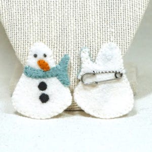 May include: Two white felt snowman pins. One pin shows the front of the snowman with a blue scarf, orange nose, and black button eyes and mouth. The other pin shows the back with a safety pin attached. The pins are on a white surface.