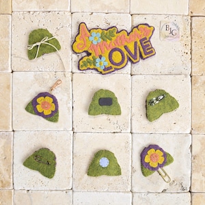 May include: A collection of handmade felt accessories, including a magnet, a pin, a paperclip, and a keychain, all in shades of green, purple, yellow, and blue. The word "Summer Love" is also featured in a felt design.