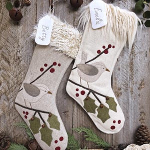 May include: Two beige fabric Christmas stockings featuring embroidered bird and holly berry designs. Each stocking has a white faux fur cuff and a white fabric tag with the names Betty and Emelia handwritten in black ink, hanging from a wooden branch.