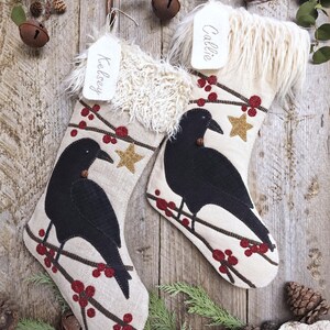 May include: Two cream-colored fabric Christmas stockings featuring black crow appliqués perched on brown branches with red berries and gold stars. Each stocking has a white faux fur cuff and a white tag with the names Kelsey and Callie written in script.