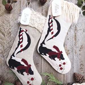 May include: Two beige fabric Christmas stockings featuring a black and white skunk wearing a red scarf, a red and white candy cane, and red berry sprigs. Each stocking has a white faux fur cuff and a white tag labeled Betty or Olivia.