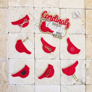 May include: A collection of red cardinal bird ornaments and pins. One ornament has a string for hanging. One pin has the text "Cardinals appear when angels are near". The items are displayed on a square tile surface.