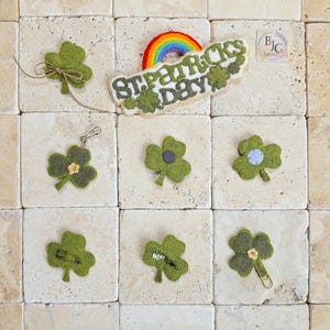 May include: A collection of green felt shamrocks and a St. Patrick's Day banner with a rainbow. Some shamrocks have pins, a magnet, or a paper clip attached. One shamrock is tied with twine. The banner reads "St. Patrick's Day".