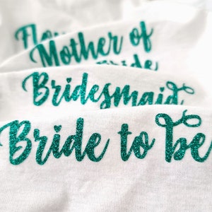 Może przedstawiać: Białe koszulki z zielonym brokatowym napisem. Koszulki mają napis "Bride to Be", "Bridesmaid" i "Mother of the Bride".