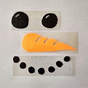 Könnte beinhalten: Drei separate Teile für ein Schneemann-Gesicht. Zwei schwarze, glitzernde Kreise für die Augen, eine orangefarbene Karottennase und eine gebogene Reihe schwarzer Punkte für den Mund. Alle Teile befinden sich auf rechteckigem, transparentem Träger.