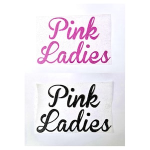 Könnte beinhalten: Zwei rechteckige Aufkleber mit dem Text "Pink Ladies". Der obere Aufkleber hat rosa Glitzertext, und der untere Aufkleber hat schwarzen Text. Beide Aufkleber befinden sich auf weißem Hintergrund.