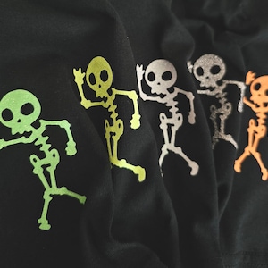 Puede incluir: Cuatro camisetas negras con diseños de esqueletos que brillan en la oscuridad en verde, plata, blanco y naranja.