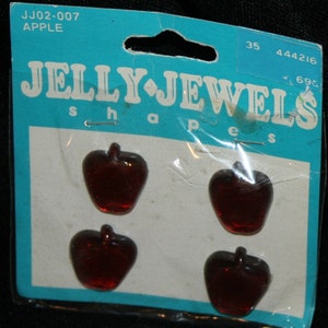 Può includere: Confezione di quattro gioielli di gelatina a forma di mela in vetro rosso. La confezione è blu e bianca e dice "JELLY JEWELS shapes".