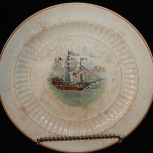 May include: A white ceramic plate with a raised rim and a decorative design of a ship with red and white sails. The ship is sailing on a blue sea with green waves.