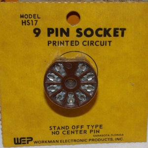 Puede incluir: Un zócalo de 9 pines marrón para circuitos impresos. El zócalo es de tipo de separación sin pin central. El embalaje es amarillo con texto negro que dice "MODEL HS17 9 PIN SOCKET PRINTED CIRCUIT STAND OFF TYPE NO CENTER PIN WEP WORKMAN ELECTRONIC PRODUCTS, INC. SARASOTA, FLORIDA".