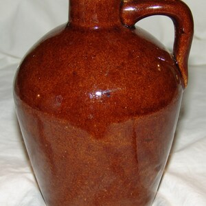 May include: A brown glazed stoneware jug with a handle. The jug has a rounded body and a small opening at the top.