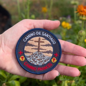 May include: A round patch with the text "CAMINO DE SANTIAGO" and "CAMINO FRANCES" in white lettering. The patch features a cross on a rocky hill, with a blue border and red and yellow accents.