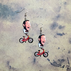 Könnte beinhalten: Ohrringe mit einem Cartoon-Bild eines Mannes mit überraschtem Gesichtsausdruck, kombiniert mit einem roten Fahrrad. Die Ohrringe haben silberfarbene Haken. Der Hintergrund ist ein meliertes Grau.