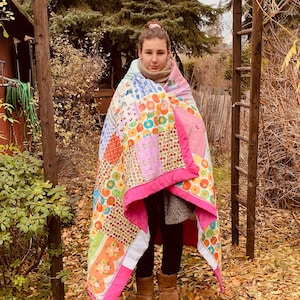 Könnte beinhalten: Eine junge Person trägt eine bunte Patchwork-Decke als Decke, die in einem Garten mit heruntergefallenen Blättern steht. Die Decke hat einen rosa Rand und zeigt verschiedene florale und geometrische Muster.
