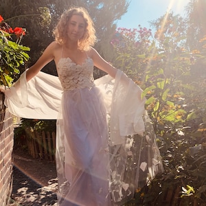 Könnte beinhalten: Eine Frau in einem weißen Spitzenkleid mit einem durchsichtigen Überwurf und einem weißen Schal. Sie steht in einem Garten mit grünem Laub und Blumen.