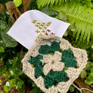 May include: A white square handkerchief with embroidered birds in shades of green and brown, next to a crocheted square with green and beige yarn. The items are set against a backdrop of green foliage.