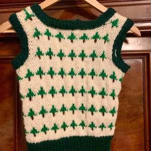 May include: A white sleeveless knitted vest with a green and brown Christmas tree pattern. The vest has a green trim around the neck and armholes.