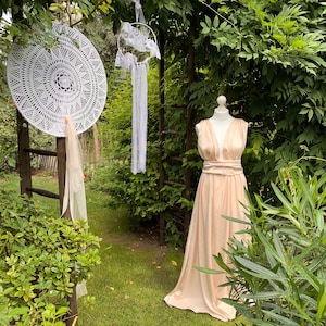 Könnte beinhalten: Ein pfirsichfarbenes Satin-Hochzeitskleid mit V-Ausschnitt und einer Schleife in der Taille. Das Kleid wird auf einer Schaufensterpuppe in einer Gartenlandschaft mit Grün und einem weißen Häkel-Traumfänger präsentiert.