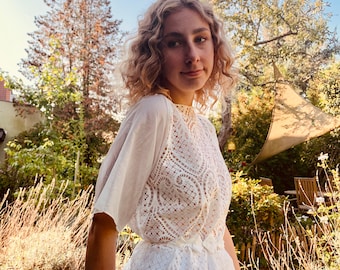May include: A woman wearing a white lace top and skirt. The top has a delicate lace pattern and a bow at the waist. The skirt is flowing and has a scalloped hem.