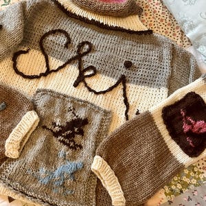May include: A brown, gray, and white knitted sweater with a turtleneck and the word "Ski" written in brown yarn across the front. The sweater has two pockets on the front, each with a different design. One pocket has a brown and blue design, and the other has a brown and pink design.