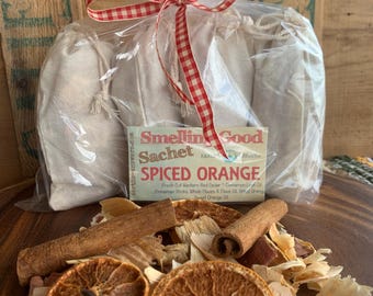 May include: A set of three white fabric sachets tied with a red and white gingham ribbon. The sachets are labeled "Smellin Good Sachet Spiced Orange" and contain a blend of spices including cinnamon sticks, cloves, and orange peel.