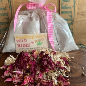 May include: A set of three small white fabric bags filled with dried hemp bark and rose petals. The bags are tied with a pink and white gingham ribbon and packaged in a clear plastic bag. The bags are labeled "Smelling Good Wild Hemp + Rose" and "Fresh Cut Dried Hemp Bark, Rose Petals, Essential Oils".