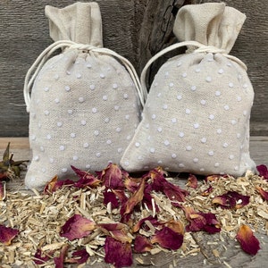 May include: Two white linen drawstring bags with white polka dots, filled with dried rose petals and wood shavings. The bags are sitting on a wooden surface.