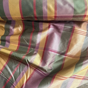 May include: A bolt of fabric featuring a plaid pattern with vertical and horizontal stripes in shades of mustard yellow, deep forest green, muted purple, and thin red lines. The material has a smooth, slightly reflective surface texture.