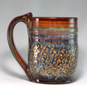 May include: A handcrafted ceramic mug with a textured surface and a unique handle design. The mug features a blend of colors, including brown, blue, and gold, creating a visually appealing aesthetic. This mug is perfect for enjoying your favorite hot beverages.
