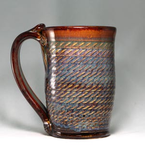 May include: A ceramic mug with a glossy brown rim and a textured body featuring a repeating diagonal pattern in shades of blue, bronze, and copper. The mug has a large, curved handle attached to the side, with a smooth, dark brown glazed finish.