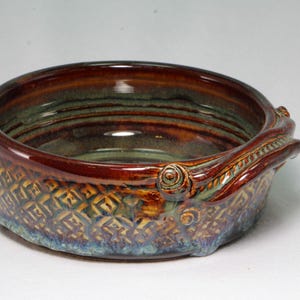 May include: A round ceramic bowl with a glossy brown and deep amber glaze. The exterior features a textured, repeating diamond pattern and a sculpted, swirling decorative element on the side. The interior is smooth with visible concentric rings.