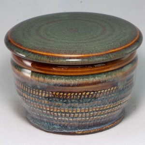 May include: A round ceramic butter crock with a lid, featuring a glossy finish in shades of deep forest green, earthy brown, and hints of blue. The exterior displays a textured, horizontal band pattern around the center of the vessel body.