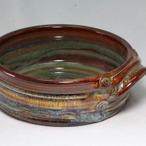 May include: A round ceramic bowl featuring a glossy glaze with blended tones of deep brown, earthy green, and hints of blue. The bowl has a thick rim and a small, integrated side handle with circular detailing, showcasing a smooth, reflective finish.