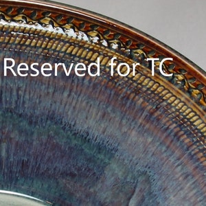 May include: A close-up of a ceramic bowl with a glossy, iridescent finish. The bowl features a dark brown rim with a decorative leaf pattern. The interior transitions from blue to purple hues, with the text "Reserved for TC" in white.