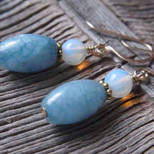 Peut inclure: Une paire de boucles d'oreilles avec des pierres ovales bleues et blanches. Les pierres bleues ont un motif marbré et les pierres blanches sont irisées. Les boucles d'oreilles sont en métal doré.