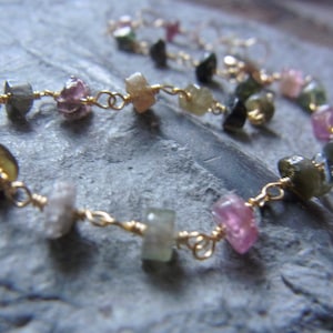 May include: A gold chain bracelet with pink, green, and black gemstones. The gemstones are faceted and have a natural, rough look.