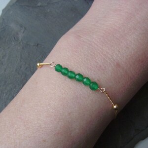 May include: A delicate gold chain bracelet with a row of green gemstone beads. The bracelet is adjustable with a sliding bead.