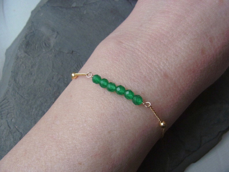 May include: A delicate gold chain bracelet with a row of green gemstone beads. The bracelet is adjustable with a sliding bead.