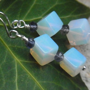 Peut inclure: Boucles d'oreilles pendantes en argent avec des accents d'opale bleue et de pierres précieuses noires. Les boucles d'oreilles ont une forme carrée et un design pendentif.
