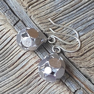 Könnte beinhalten: Ein Paar silberfarbene Ohrhänger. Jeder Ohrring hat ein facettiertes, kugelförmiges Design mit einer kleinen, kreisförmigen Öffnung. Die Ohrringe hängen an gebogenen, silberfarbenen Ohrhaken. Der Hintergrund ist eine verwitterte Holzoberfläche.