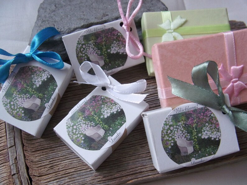 May include: Five small white boxes with a floral design and the text "Blickfang!" and "Danke". Each box is tied with a different colored ribbon: blue, white, pink, green, and pink.
