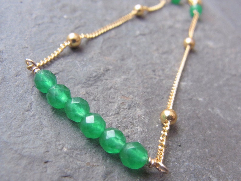 May include: A gold chain bracelet with a green gemstone slider. The bracelet features a delicate chain and small gold beads.
