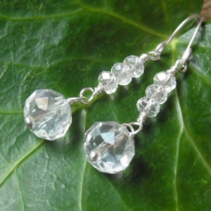 May include: A pair of silver earrings with clear crystal beads. The earrings are dangling and have a teardrop-shaped crystal at the bottom.