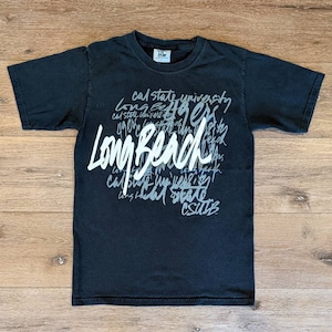 Könnte beinhalten: Schwarzes T-Shirt mit dem Schriftzug "Long Beach" in großem, weißem Schriftzug, umgeben von kleinerem Text mit "Cal State University" und "CSULB". Das T-Shirt liegt flach auf einer Holzoberfläche.