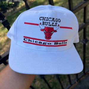 May include: White baseball cap with the Chicago Bulls logo embroidered on the front. The logo is red and black with a red and white stripe above and below it. The text "Chicago Bulls" is embroidered in black below the logo.