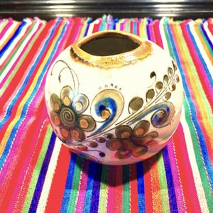 Peut inclure: Un vase en céramique blanche de forme ronde avec une large ouverture. Le vase est décoré de motifs floraux marrons, bleus et dorés.