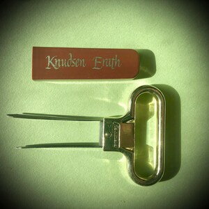 Puede incluir: Un abridor de botellas dorado con una caja de madera grabada con el texto "Knudsen Eraeth".