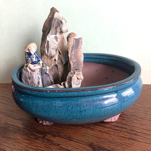 May include: A teal ceramic bonsai pot with a miniature zen garden scene. The garden features a rock formation and a small figurine of a person in a hat. The pot is round with a glossy finish and sits on small feet.