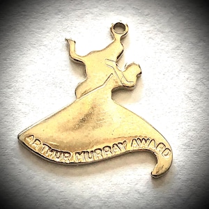 Puede incluir: Un dije de metal en tono dorado con la forma de una pareja bailando en una pose giratoria. La base del dije presenta el texto en relieve ARTHUR MURRAY AWARD. En la parte superior hay un pequeño aro circular para sujetarlo a una cadena.